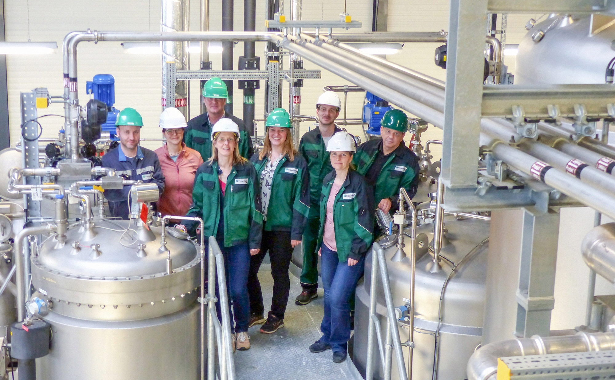 Gruppenbild von Mitarbeitenden in grüner Arbeitskleidung und Helmen der Gruppe »Bioprozess-Skalierung« am Fraunhofer CBP in einem Technikum, auf einer Anlage. 